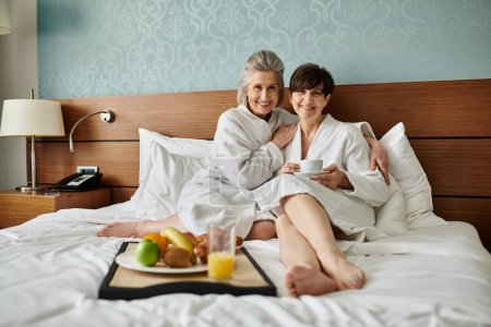 Tender senior lesbian couple sitting together on the edge of a bed with love and comfort.