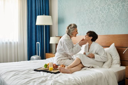 Photo for Two senior women, a loving lesbian couple, sit peacefully on top of a bed. - Royalty Free Image