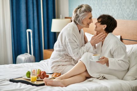 A senior lesbian couple sits tenderly on a bed.