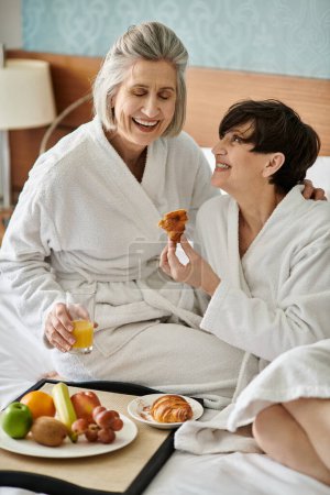 Two senior lesbian women lovingly embrace while sitting on a bed in a hotel room.