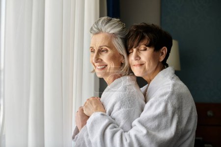 Photo for Senior lesbian couple embrace warmly in a hotel room. - Royalty Free Image