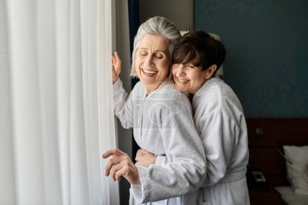 Two senior lesbian women share a tender hug in front of a window.
