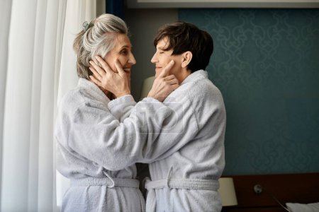 Photo for Senior lesbian couple in bathrobes admire the view through a window. - Royalty Free Image