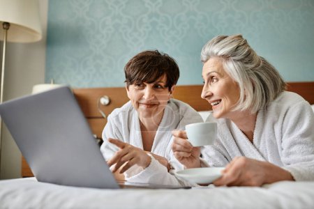 Zwei Frauen, ein älteres lesbisches Paar, sitzen auf dem Bett, vertieft mit Laptop-Bildschirm in zartem Moment.