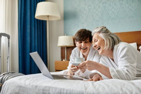 Ältere lesbische Paar sitzt auf einem Bett, konzentriert auf einem Laptop-Bildschirm.