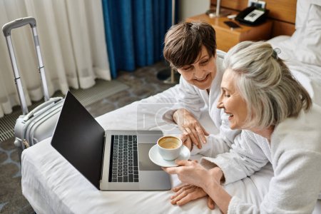 Ein älteres lesbisches Paar sitzt auf einem Bett und benutzt einen Laptop.