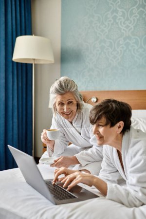 A senior lesbian couple peacefully lounging in bed, engrossed in a laptop.
