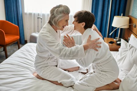 Téléchargez les photos : Deux femmes âgées profitant d'un moment tendre, assises sur un lit luxueux dans un hôtel. - en image libre de droit