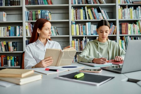 Foto de Una tutora pelirroja y una adolescente se sientan en la mesa de la biblioteca. El tutor enseña mientras usa una computadora portátil para la educación moderna. - Imagen libre de derechos