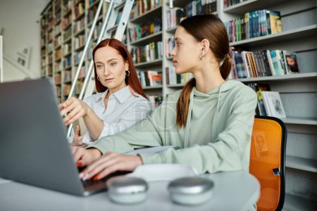Téléchargez les photos : Un tuteur aux cheveux roux guide un étudiant dans une bibliothèque, tous deux travaillant sur un ordinateur portable pour des cours après l'école. L'éducation moderne en action. - en image libre de droit