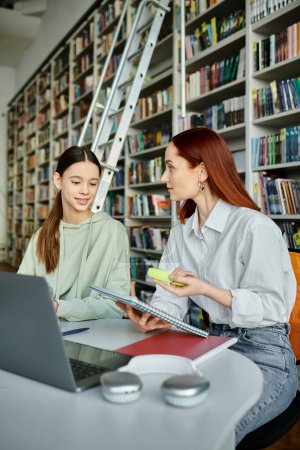 Redhead tutor teaches teenage girl in library using laptop for modern education after school lessons.