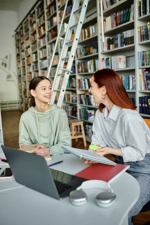Photo for A tutor with red hair teaches a teenage girl in a library, using a laptop for after-school lessons on modern education. - Royalty Free Image