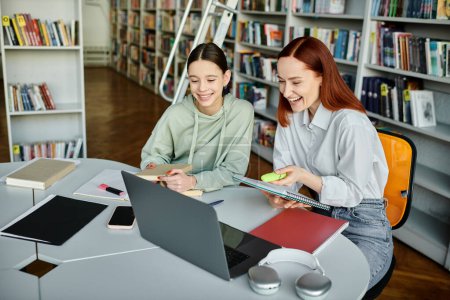 Eine rothaarige Tutorin unterrichtet ein Teenager-Mädchen in einer Bibliothek und nimmt am Nachmittagsunterricht mit einem Laptop teil..