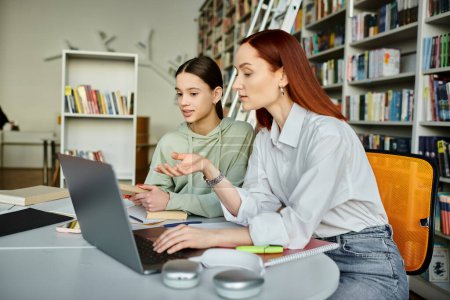 Foto de Un tutor con cabello rojo guía a una adolescente durante una lección después de la escuela usando una computadora portátil en un entorno de biblioteca. - Imagen libre de derechos