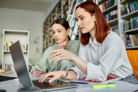 Tuteur rousse et adolescente engagés dans des leçons après l'école, en utilisant un ordinateur portable pour l'éducation moderne dans un cadre de bibliothèque.