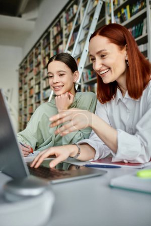 Un tuteur roux enseigne à une adolescente dans une bibliothèque, travaillant tous deux intensément sur un ordinateur portable pour l'éducation moderne après l'école.