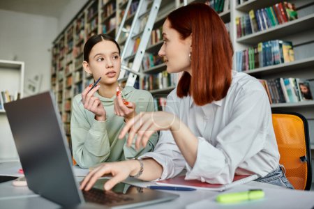 Photo for A redhead tutor teaches a teenage girl in a library, both engrossed in a laptop during an after-school lesson. - Royalty Free Image