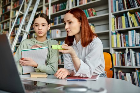Teenager Mädchen und Tutor mit roten Haaren, engagieren sich in einer modernen Bildungsveranstaltung in einer Bibliothek, die sich auf einen Laptop-Bildschirm konzentriert.