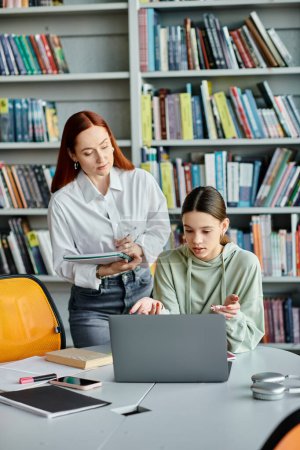 Redhead tutor and teenage girl engage in modern education, discussing school lessons on a laptop in a library setting.