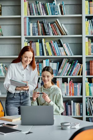 Eine rothaarige Tutorin unterrichtet ein Teenager-Mädchen in einer Bibliothek, beide vertieft mit einem Laptop während des After-School-Unterrichts.