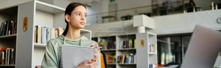 Ein Mädchen im Teenageralter steht in einer Bibliothek vor einem Bücherregal, vertieft in das Lernen und Hausaufgaben machen auf ihrem Laptop nach der Schule.