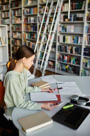 Téléchargez les photos : Une adolescente immergée dans ses études, travaillant diligemment sur un ordinateur portable à une table dans une bibliothèque calme. - en image libre de droit