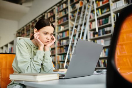 Une adolescente avec une expression paisible est assise à un bureau dans une bibliothèque, se concentrant sur son ordinateur portable pendant qu'elle fait du travail scolaire.