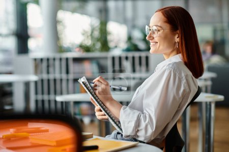 Photo for A red-haired woman sits at a desk, writing on a clipboard while teaching an online lesson using a laptop after school. - Royalty Free Image