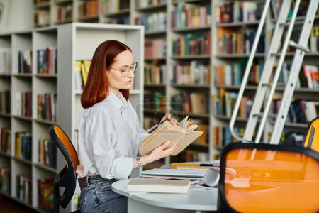 A redheaded woman sits at a table in a library, engrossed in reading a book.