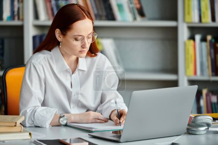 Photo for A redhead female tutor focuses on teaching an after school lesson while working on a laptop in a library. - Royalty Free Image