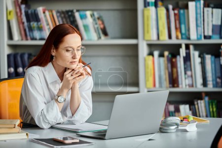 Photo for A redheaded female tutor is teaching an online lesson using a laptop at a desk. - Royalty Free Image