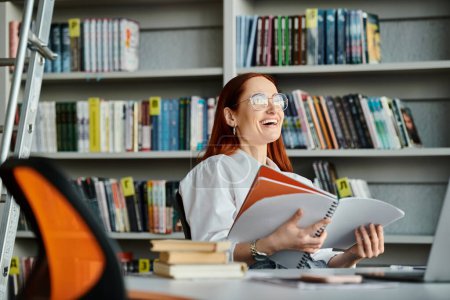 Téléchargez les photos : Une tutrice rousse enseigne en ligne, assise à un bureau de bibliothèque absorbé par la lecture d'un livre après sa leçon après l'école. - en image libre de droit