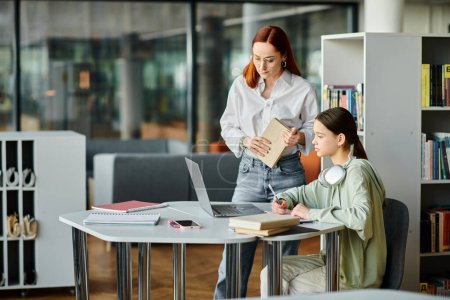 Una mujer pelirroja es tutora de una adolescente en una biblioteca, colaborando en una computadora portátil para lecciones extraescolares.