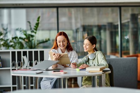 Photo for A redhead woman is tutoring a teenage girl at a table in an office, using a laptop for modern education after school lessons. - Royalty Free Image