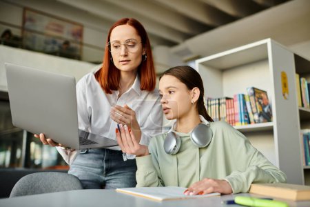 Photo for Redheaded woman tutors a teenage girl in a library, both engaged and focused on a laptop screen for modern education. - Royalty Free Image