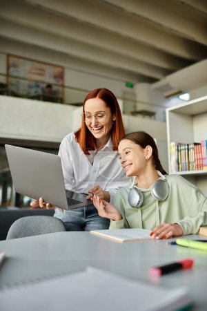 Foto de Mujer pelirroja tutores una adolescente en una biblioteca, ambos centrados en un ordenador portátil, participan en lecciones después de la escuela. - Imagen libre de derechos