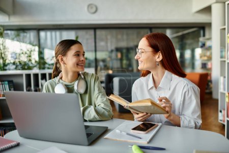 Photo for A redhead woman is teaching a teenage girl at a table while both are focused on a laptop screen. - Royalty Free Image
