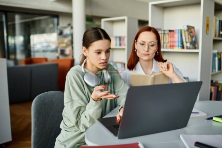 Téléchargez les photos : Une rousse enseigne à une adolescente à une table, toutes deux concentrées sur un ordinateur portable pendant une leçon après l'école. - en image libre de droit