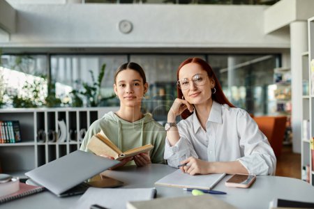 Photo for A redhead woman is tutoring a teenage girl, both engrossed in after-school studies at a table in a library. - Royalty Free Image