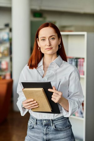 Photo for A redheaded woman holds a book in a library, embodying modern education. - Royalty Free Image