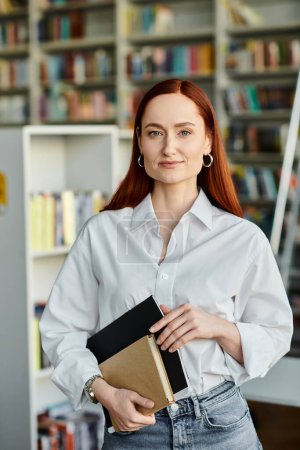 Photo for In a library, a redhead woman holds a book, after-school lessons. - Royalty Free Image