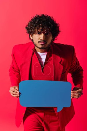 Schöner junger indischer Mann im leuchtend roten Anzug hält eine Sprechblase vor einem lebhaften Hintergrund.