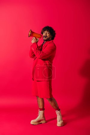 A young Indian man in a vibrant red suit holding a red megaphone.