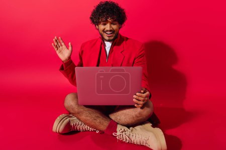 A man in vibrant attire, seated on floor, engrossed in laptop work.