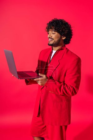 Handsome young Indian man in a vibrant red suit holds a laptop.