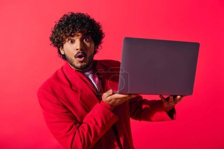 A stylish young Indian man in a red jacket holding a laptop.