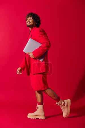 A stylish young Indian man in a striking red suit holding a laptop.