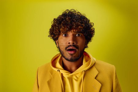 Handsome young Indian man with curly hair posing in a vibrant yellow jacket on a vivid backdrop.
