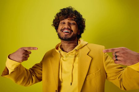 Ein gutaussehender junger indischer Mann in einer gelben Jacke zeigt auf etwas vor einer lebhaften Kulisse.