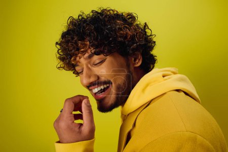 A handsome young Indian man with curly hair striking a pose in a vibrant yellow jacket on a vivid backdrop.